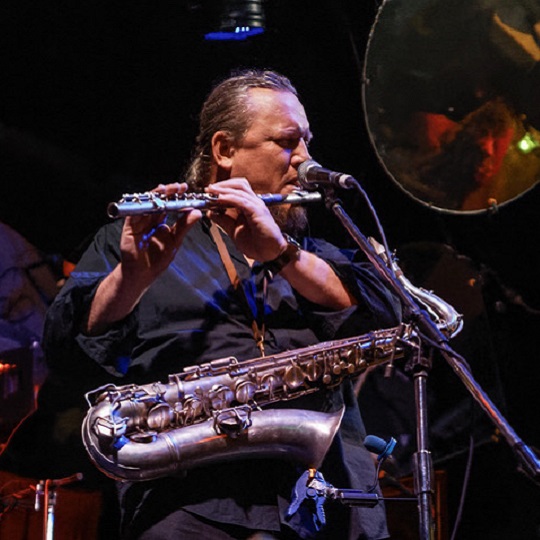 James Ryan sits on stage in black shirt and jeans, his hair is in a ponytail an he has a saxophone on his lap and a flute pursed to his lips, there is lighting on James